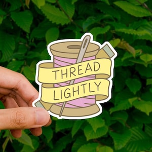 May include: A die-cut sticker featuring a wooden spool of light pink thread with a silver sewing needle inserted through the center. A yellow banner wraps around the spool with the black text THREAD LIGHTLY printed in a clear, bold font.