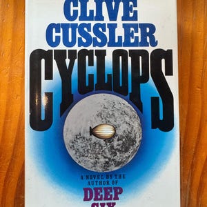 Puede incluir: Una portada de libro titulada Cyclops de Clive Cussler con un fondo degradado en azul y blanco. La portada presenta un gráfico grande de una luna gris texturizada con una pequeña ilustración de un dirigible en el centro y el texto A NOVEL BY THE AUTHOR OF DEEP SIX.