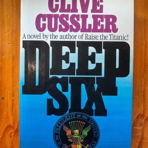Puede incluir: Un libro de bolsillo titulado Deep Six de Clive Cussler. La portada presenta el nombre del autor en letras grandes de color púrpura, el título en negro y un fondo con degradado azul. Un sello circular del Presidente de EE. UU. está centrado.