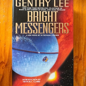 Può includere: Un libro con copertina rigida intitolato Bright Messengers di Gentry Lee. La copertina presenta un grande oggetto sferico blu nello spazio, una piccola navicella spaziale e una superficie planetaria arancione brillante. Il testo include coautore di Rama II.