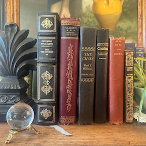 Pode incluir: Uma coleção de livros antigos de capa dura, incluindo The Decameron, Tolerance, The Chain, Catalina, Jane Eyre e Catherine, dispostos ao lado de um remate decorativo preto, uma esfera de vidro transparente num suporte dourado e uma planta.