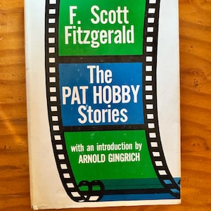 May include: A book titled The Pat Hobby Stories by F. Scott Fitzgerald with an introduction by Arnold Gingrich. The cover features a graphic design resembling a black and white film strip framing green and blue rectangular text blocks.