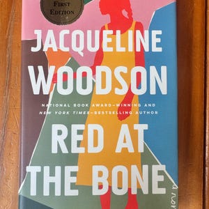 Pode incluir: Um livro de capa dura intitulado Red at the Bone de Jacqueline Woodson. A capa apresenta a silhueta vermelha de uma pessoa com um vestido amarelo sobre um fundo geométrico azul, verde e rosa. Um adesivo circular diz Signed First Edition.