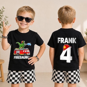 May include: A black t-shirt featuring a green dinosaur wearing a red firefighter helmet on a red fire truck with the text FIRESAURUS on the front and the name FRANK with the number 4 and a red helmet graphic on the back, paired with checkered shorts.