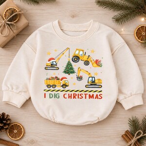 May include: A cream-colored long-sleeve sweatshirt featuring a graphic print of yellow construction vehicles, a Christmas tree, and wrapped gifts. The text "I DIG CHRISTMAS" is printed in green and red letters below the construction scene.
