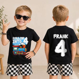 May include: A black short-sleeved t-shirt featuring a colorful train graphic with the text Rollin' Into 4 on the front and the name Frank with the number 4 on the back, paired with black and white checkered shorts. The design includes blue and red.