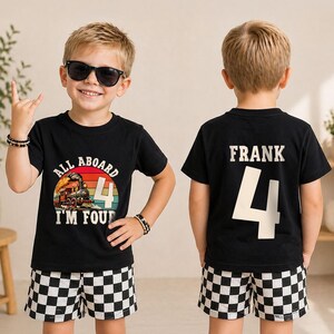 May include: A black t-shirt featuring a graphic of a steam train with the text ALL ABOARD I'M FOUR and a large number 4 on the front, and the name FRANK with a large number 4 on the back, paired with black and white checkered shorts.