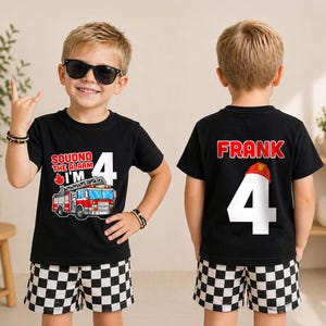 May include: A black t-shirt featuring a graphic of a fire truck, the text "SOUDND THE ALARM I'M 4" on the front, and the name "FRANK" with the number "4" and a red fire helmet graphic on the back, paired with black and white checkered shorts.