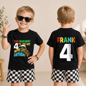 May include: A black short-sleeved t-shirt featuring a colorful train graphic with the text THE RAILWAY 4TH TO MY BIRTHDAY on the front and the name FRANK with the number 4 in large white font on the back, paired with black and white checkered shorts.