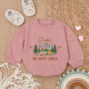 May include: A dusty rose baby sweatshirt with long sleeves. The front features a camping scene with a tent, mountains, and trees, along with the text "Jordan EST. 2024 ONE HAPPY CAMPER". Two pairs of baby shoes are in the background.