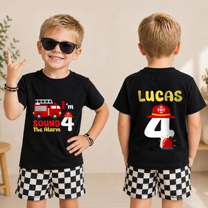 May include: A black short-sleeved t-shirt featuring a fire truck graphic, the text 'I'm 4 Sound the Alarm' on the front, and the name 'LUCAS' with a large number 4 and a firefighter helmet graphic on the back, paired with black and white checkered shorts.
