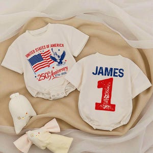 May include: Two white baby onesies. One features the text "UNITED STATES OF AMERICA" with an American flag and an eagle graphic. The other onesie has the name "JAMES" and the number "1" printed on it. Soft toys are in the foreground.