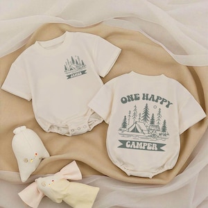 May include: Two off-white baby rompers with short sleeves. One has the name "JAMES" and a camping scene printed in teal. The other says "ONE HAPPY CAMPER" with a camping scene. Two stuffed toys are in the foreground.