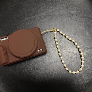 May include: A brown Canon G7X digital camera with a lens cap attached to a wrist strap made of alternating white faux pearls and small gold-toned beads. The camera body features a textured grip on the left side and the brand name printed in white.