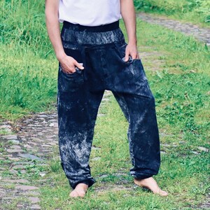 Könnte beinhalten: Eine Person trägt ein weißes T-Shirt und eine schwarze und weiße gemusterte Hose. Die Person steht auf einem Weg mit grünem Gras und Steinen.