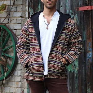 Può includere: Un uomo indossa una giacca a motivi marroni e neri con una camicia bianca sotto. Indossa pantaloni di velluto a coste marroni e ha una collana con un ciondolo.