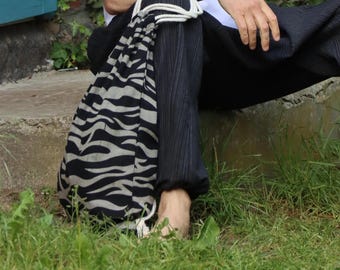 May include: A man wearing a white t-shirt with a small logo on the left chest, black pants, and a zebra-print drawstring bag. He is sitting on a concrete step with green grass in the background.