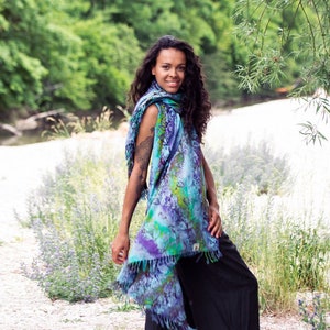 May include: A woman wearing a blue, green, and purple patterned scarf with fringe. She is standing in a field of flowers and looking at the camera.