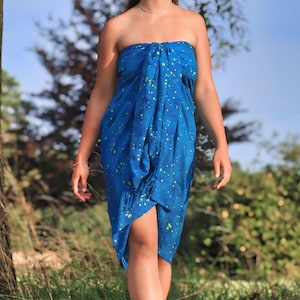 May include: A woman wearing a blue and green patterned sarong. The sarong is tied at the waist and has a loose fit. The woman is standing in a grassy field.