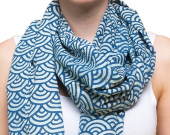 May include: A blue and white patterned scarf with a wave design. The scarf is worn around the neck of a person wearing a white tank top and blue jeans.