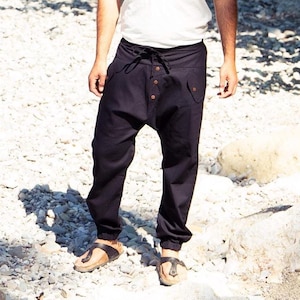 Op de afbeelding: Een persoon met een wit t-shirt, zwarte broek en sandalen staat op een rotsachtig oppervlak. De persoon draagt een zonnebril en heeft een baard. De achtergrond omvat groen gebladerte en zonlicht.
