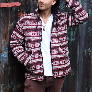 Pode incluir: Um homem vestindo uma jaqueta com capuz vermelho e branco com um padrão e fecho de correr. Ele está usando calças marrons e uma camiseta branca. Ele está em pé em frente a uma porta de madeira.