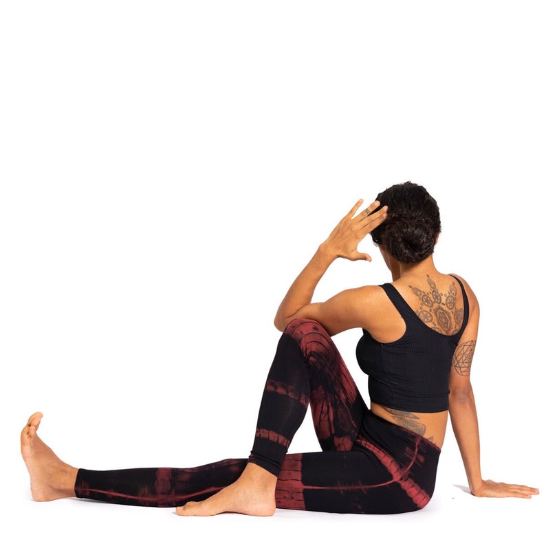 May include: A person is in a seated yoga pose, wearing a black ribbed crop top and black leggings with a red tie-dye pattern. The person's arm is raised, with the hand near the head. A tattoo is visible on the back.