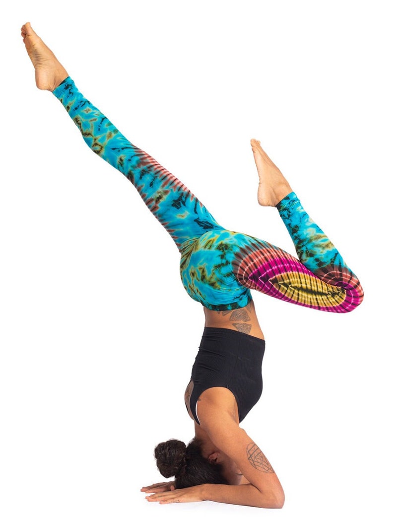 May include: A person in a blue, green, pink, and yellow tie-dye leggings is doing a headstand. They are wearing a black tank top and have their hair pulled back in a bun.