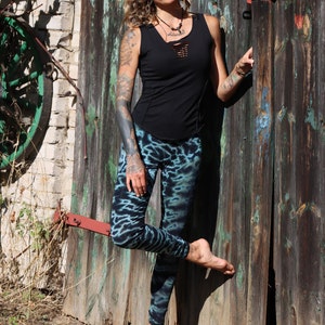 May include: A black tank top with a cut-out neckline and fitted design, paired with blue and black tie-dye leggings. The leggings have a unique pattern. The person is leaning against a weathered wooden door.