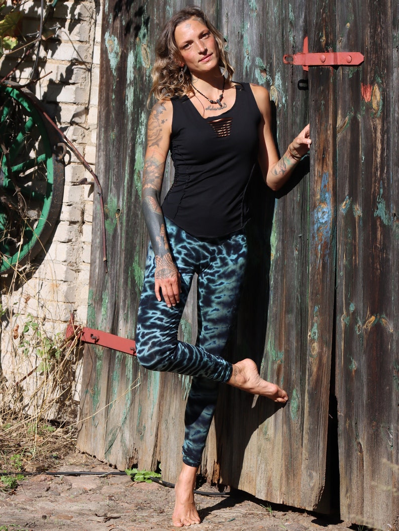 May include: A black tank top with a cut-out neckline and fitted design, paired with blue and black tie-dye leggings. The leggings have a unique pattern. The person is leaning against a weathered wooden door.