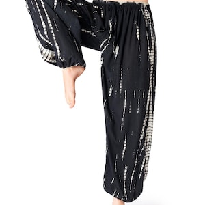 Weite schwarze Tie-Dye-Haremshose mit beigen Streifen, elastischem Bund und Knöchelbündchen, Modell hebt ein Bein im Yoga-Stil.