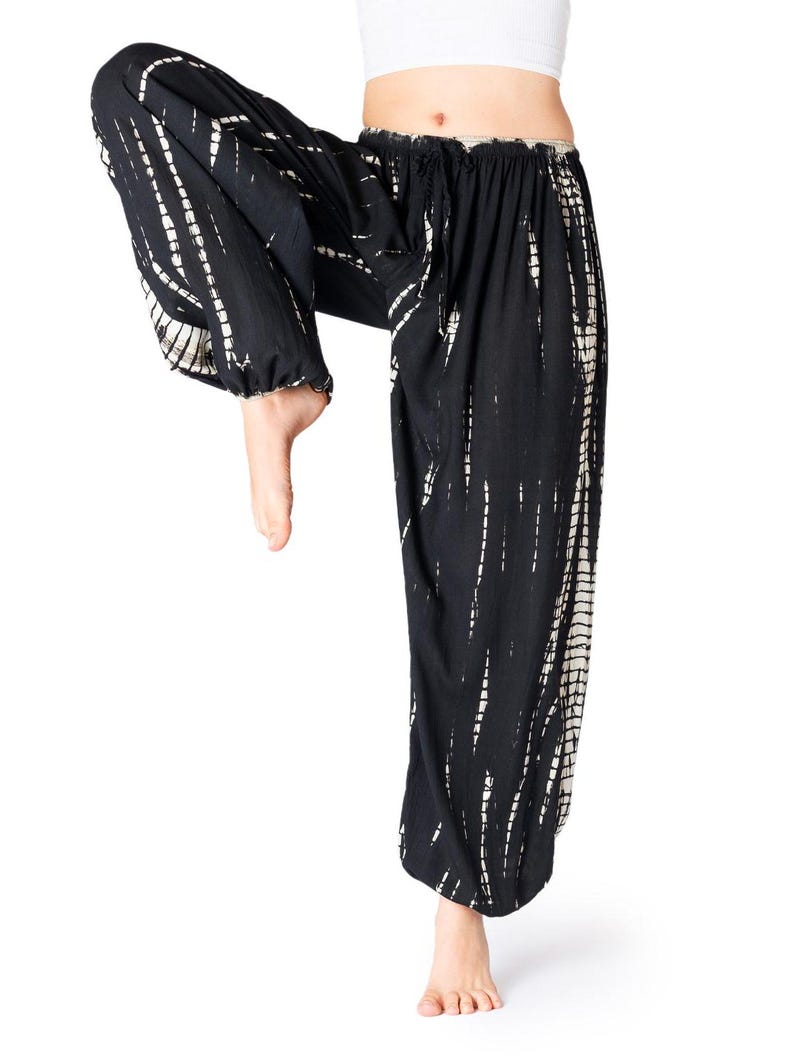Weite schwarze Tie-Dye-Haremshose mit beigen Streifen, elastischem Bund und Knöchelbündchen, Modell hebt ein Bein im Yoga-Stil.