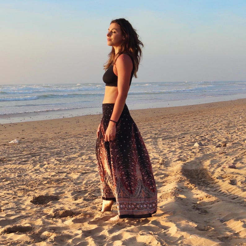 May include: A black crop top and a long, flowing skirt with a black background and a red and white paisley design. The skirt has a wide, decorative border. The person is standing on a sandy beach with the ocean in the background.