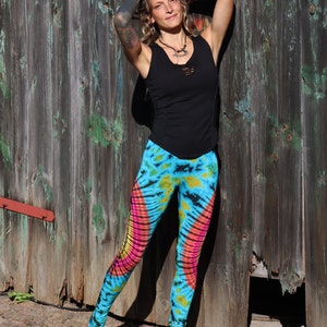 May include: A woman wearing a black tank top and tie-dye leggings with pink, yellow, and blue colors. She is standing in front of a wooden wall with a broken section.