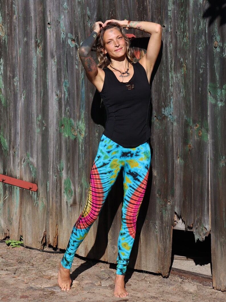 May include: A woman wearing a black tank top and tie-dye leggings with pink, yellow, and blue colors. She is standing in front of a wooden wall with a broken section.