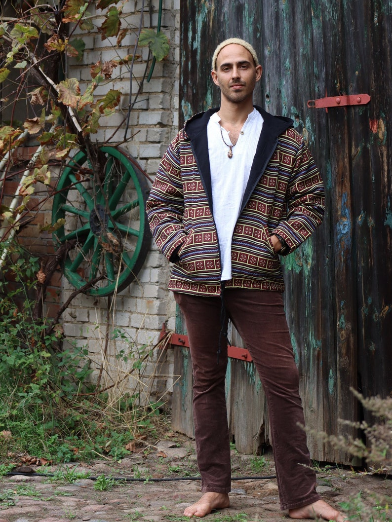 May include: A man wearing a brown and black patterned jacket with a white shirt underneath. He is wearing brown corduroy pants and has a necklace with a pendant.