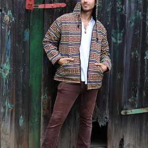 May include: A man wearing a brown, black, and yellow patterned hooded jacket with a zipper closure. He is wearing brown corduroy pants and is standing in front of a wooden door.