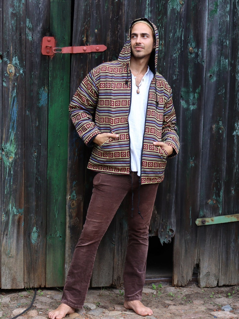 May include: A man wearing a brown, black, and yellow patterned hooded jacket with a zipper closure. He is wearing brown corduroy pants and is standing in front of a wooden door.