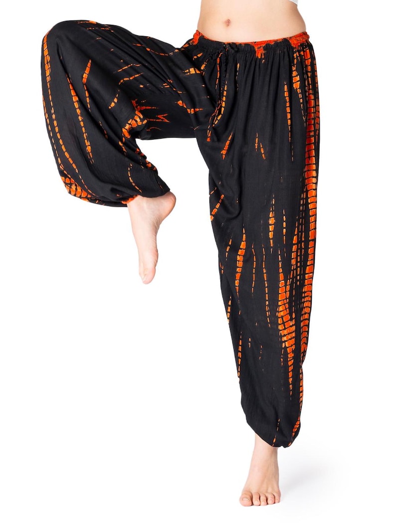 Schwarze Haremshose mit auffälligen orangefarbenen Tie-Dye-Streifen, elastischer Bund und Knöchelbündchen, Modell hebt ein Bein im Yoga-Stil.