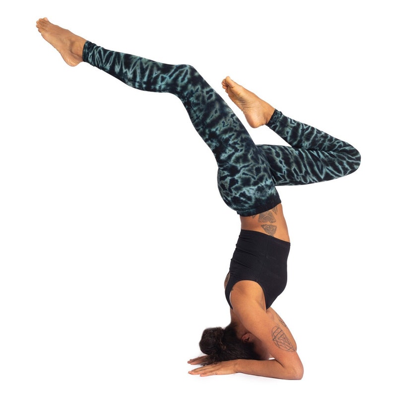 May include: Black yoga top and leggings with a teal tie-dye pattern. The leggings are full-length. The person is in a headstand pose, with legs extended. The background is white.