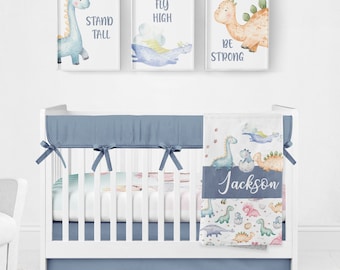 May include: A baby crib with a blue and white dinosaur themed bedding set. The crib has a blue bumper and a white sheet with a dinosaur print. The bedding set includes a blanket with the name "Jackson" printed on it. The crib is decorated with three framed prints featuring dinosaurs with the words "Stand Tall", "Fly High", and "Be Strong".
