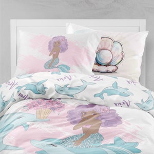 K&ouml;nnte beinhalten: Ein rosa-wei&szlig;es Bettw&auml;sche-Set mit einem Meerjungfrauen- und Delfin-Design. Das Bettw&auml;sche-Set enth&auml;lt eine Meerjungfrau mit lila Haaren und eine Muschel mit einer Perle. Das Bettw&auml;sche-Set zeigt auch den Namen "Anya", der sich im gesamten Design wiederholt.