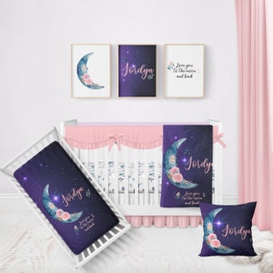 May include: A nursery bedding set with a purple and blue starry sky design featuring a crescent moon with pink flowers and the name "Jordyn". The set includes a crib sheet, a blanket, and a pillow.