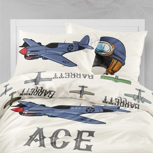May include: A white and cream colored bedding set with a blue and white airplane design. The bedding set features the word "ACE" in a large, bold font. The pillowcase features a pilot's helmet with goggles and the name "BARRETT" written backwards.