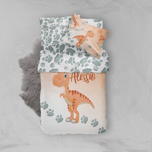 May include: A white and green dinosaur bedding set with a dinosaur footprint pattern. The duvet cover features a cartoon dinosaur with the name Alessio printed on it.