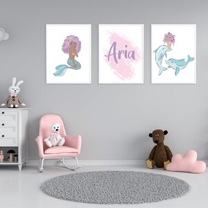 K&ouml;nnte beinhalten: Drei gerahmte Drucke an einer grauen Wand. Der linke Druck zeigt eine Meerjungfrau mit lila Haaren und einem blauen Schwanz. Der mittlere Druck hat einen rosa und lila Aquarellhintergrund mit dem Namen "Aria" in lila geschrieben. Der rechte Druck zeigt zwei Delfine, die mit einem Strau&szlig; lila Blumen schwimmen.