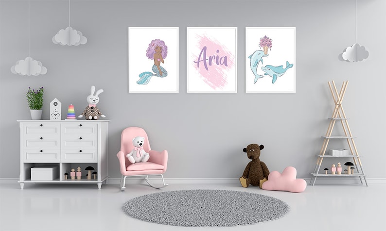 K&ouml;nnte beinhalten: Drei gerahmte Drucke an einer grauen Wand. Der linke Druck zeigt eine Meerjungfrau mit lila Haaren und einem blauen Schwanz. Der mittlere Druck hat einen rosa und lila Aquarellhintergrund mit dem Namen "Aria" in lila geschrieben. Der rechte Druck zeigt zwei Delfine, die mit einem Strau&szlig; lila Blumen schwimmen.