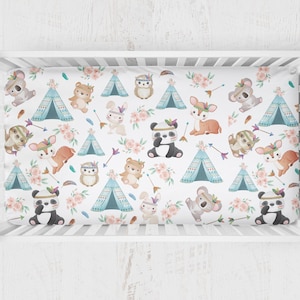 May include: A white crib sheet with a whimsical pattern featuring various animals, including deer, owls, koalas, pandas, and bunnies. The animals are wearing headdresses and are surrounded by teepees, arrows, and pink flowers.