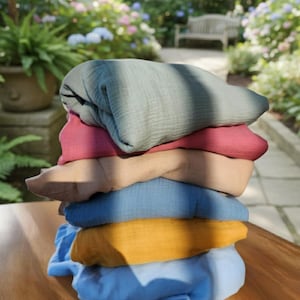 May include: A stack of folded, soft fabric items in various colors, including blue, yellow, brown, pink, and green. The items are stacked on a wooden surface, with a blurred outdoor background of greenery and flowers.