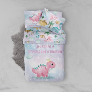 May include: A pink and blue bedding set with a dinosaur theme. The bedding features a pink dinosaur with a crown, surrounded by clouds and palm trees. The text "You can be a Princess and a Dinosaur" is printed on the bedding.